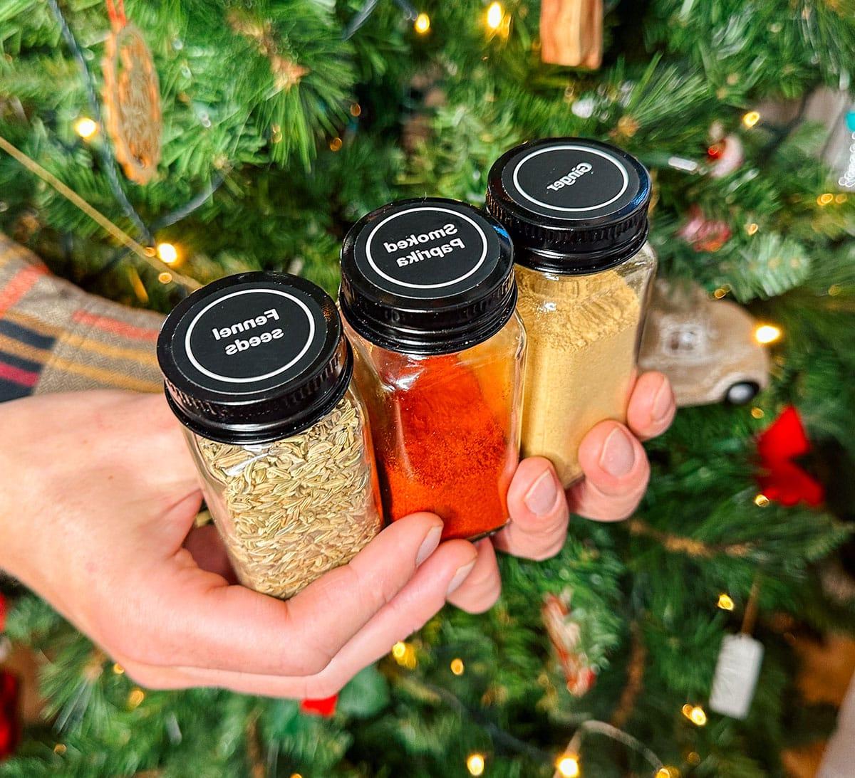47 Geschenkideen für Feinschmecker 16 Beschriftete Gewürzgläser mit passenden Messlöffeln als Geschenk vor einem Weihnachtsbaum.
