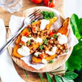 Mediterran gefüllte Süßkartoffeln, serviert als gebackene Hälften, garniert mit gerösteten Kichererbsen, gewürfelten Tomaten, Sauce und Petersilie auf einem mit Papier ausgelegten Holzbrett. Begleitet von frischen Tomaten und einem Glas – die perfekte Geschmacksmischung bei jedem Bissen.