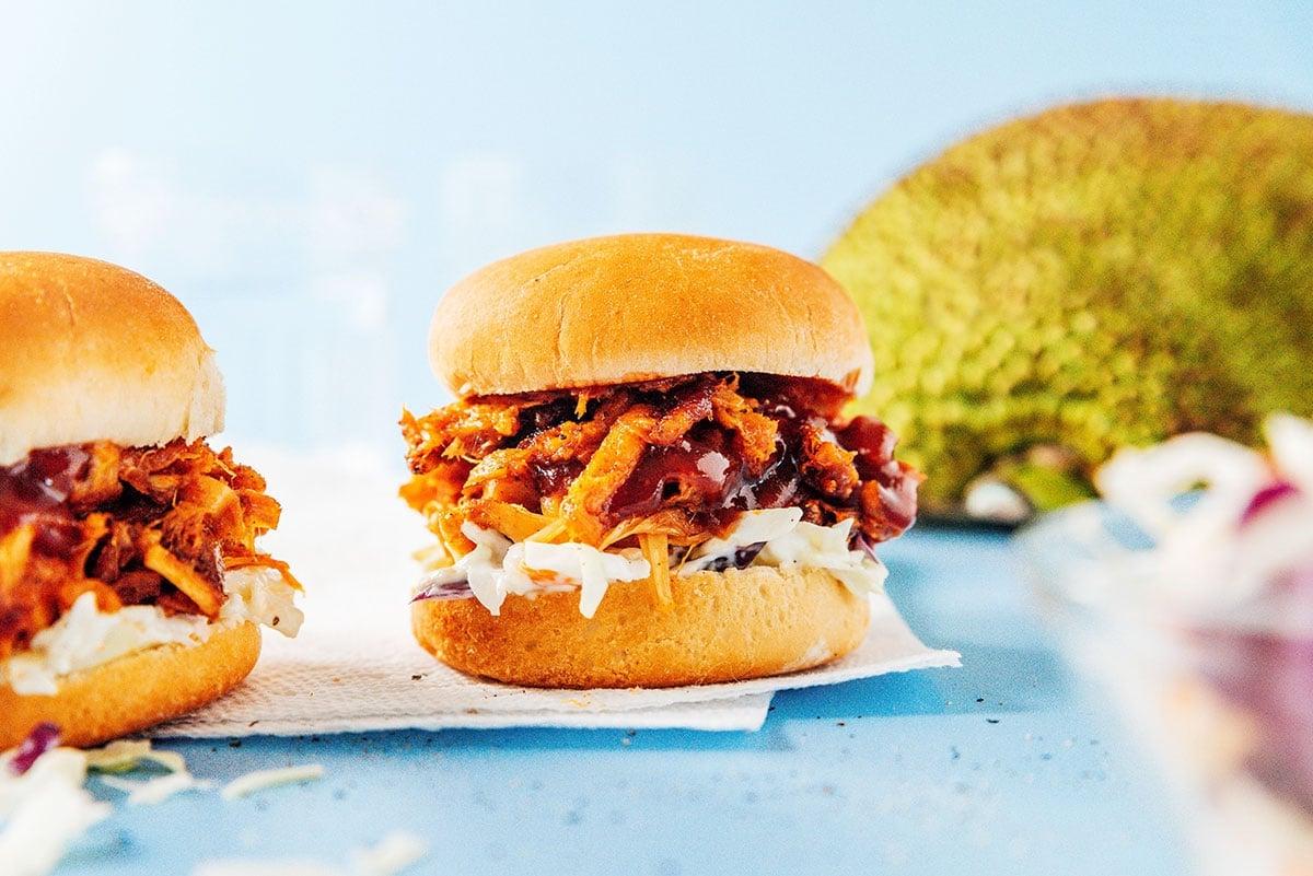 BBQ Jackfrucht Pulled Pork (Fertig in 30 Minuten) 5 Ein Detailbild eines BBQ Jackfrucht Sliders mit Krautsalat und heruntertropfender BBQ-Sauce, mit einer ganzen Jackfrucht im Hintergrund.