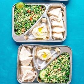 Drei Metalltabletts mit unterteilten Fächern präsentieren einen lebendigen Tabouleh-Salat, dazu cremigen, mit Olivenöl beträufelten Hummus, Pitabrot und erfrischende Gurkenscheiben, alles vor einem atemberaubenden türkisfarbenen Hintergrund.