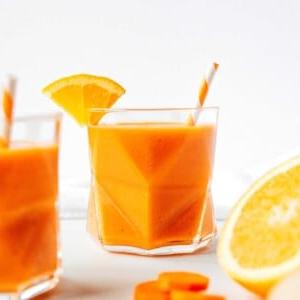 Karotten-Ingwer-Smoothie 8 Ein geometrisches Glas Karotten-Smoothie, gemixt mit leuchtender Orange, garniert mit einem frischen Orangenkeil und Strohhalm. Geschnittene Karotte und eine halbe Orange liegen verlockend im Vordergrund.