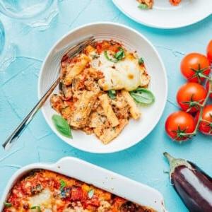 Baked vegetable ziti with cheese and herbs in a white dish, nestled beside plates of the same, fresh tomatoes, eggplant, and empty glasses on a light blue surface.