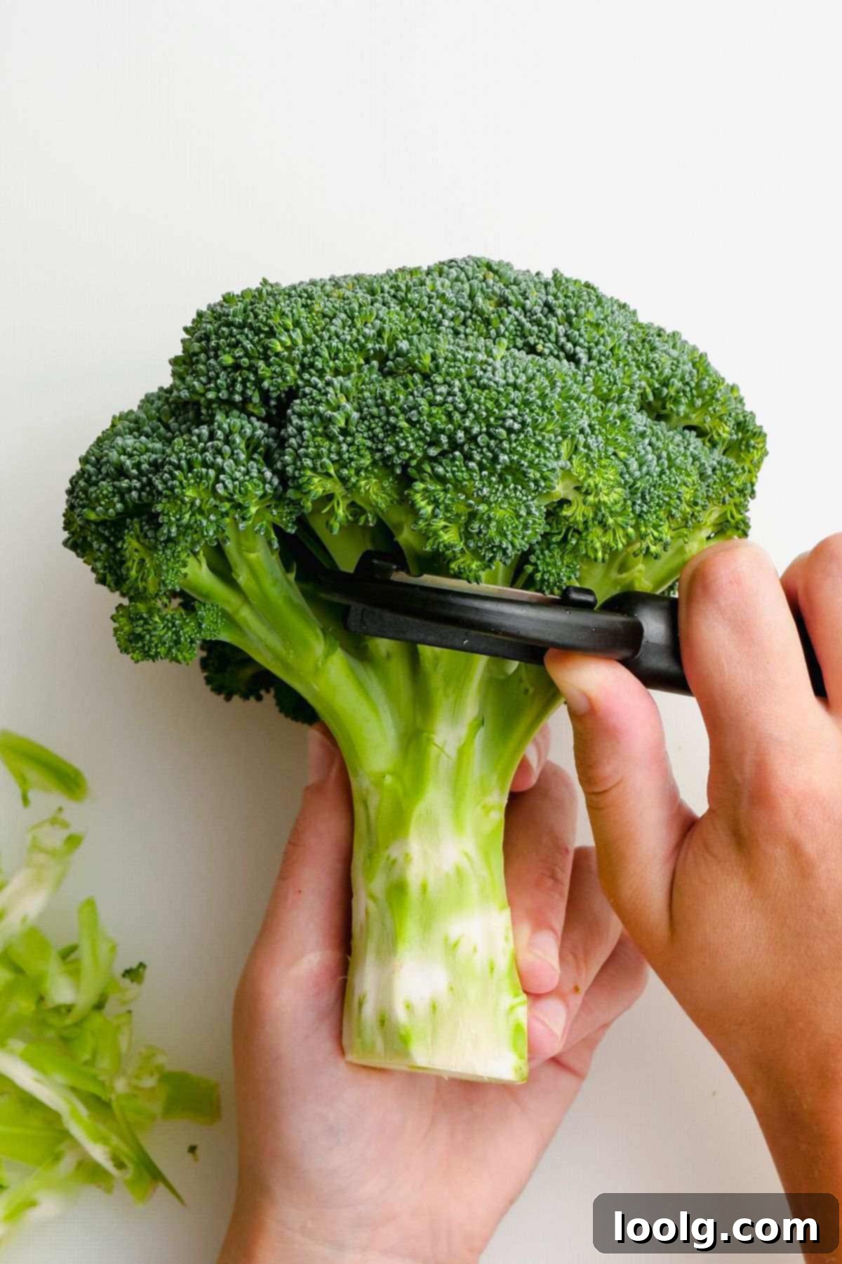 A hand peeling the fibrous outer skin off a broccoli stem with a vegetable peeler.
