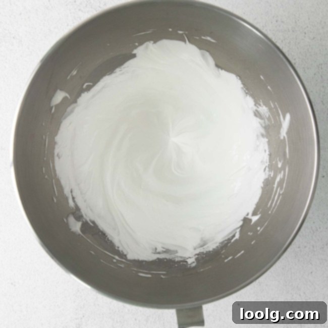 Meringue mixture with stiff peaks in a stand mixer bowl, showing the glossy and firm consistency.