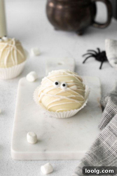 Two Halloween Hot Chocolate Bombs in a small bowl, decorated like mummies with white chocolate drizzles and candy eyes.