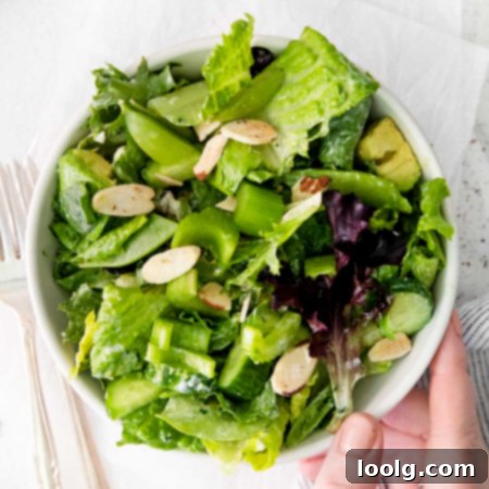 Top view close up of a hand reaching for a green goddess salad in a white bowl full of romaine hearts, mixed salad greens, cucumber, avocado, snap peas, celery, and almonds, for garnish giving it an extra crunch.