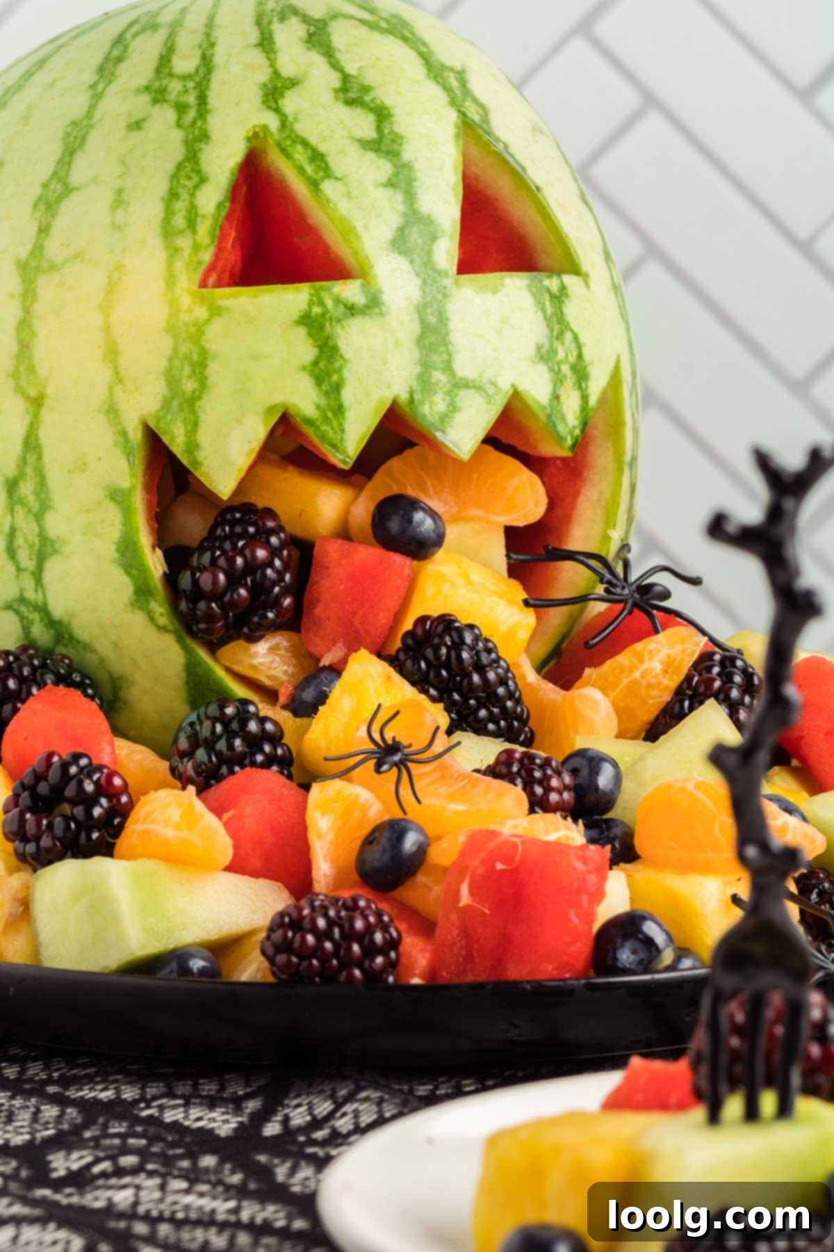 A close-up photograph of a watermelon carved into a jack-o'-lantern face, with an abundance of fresh fruit and berries playfully spilling out of its jagged mouth, adorned with a few fake spiders for a festive Halloween fruit salad presentation.