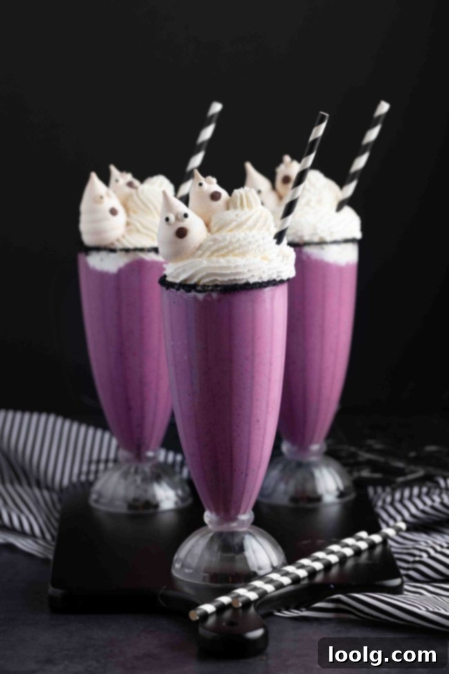 Two Halloween milkshakes with purple liquid, topped with whipped cream and meringue ghosts, in front of a spooky background.