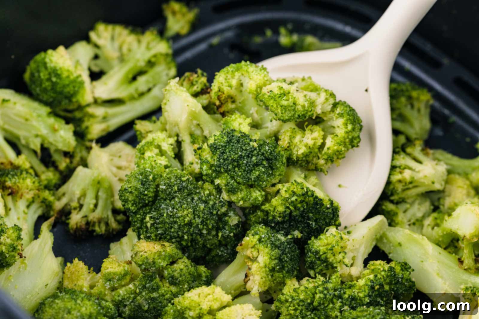 Close-up of air fryer frozen broccoli florets in a black pan, with a white plastic spoon scooping some pieces. The broccoli appears lightly seasoned and roasted.