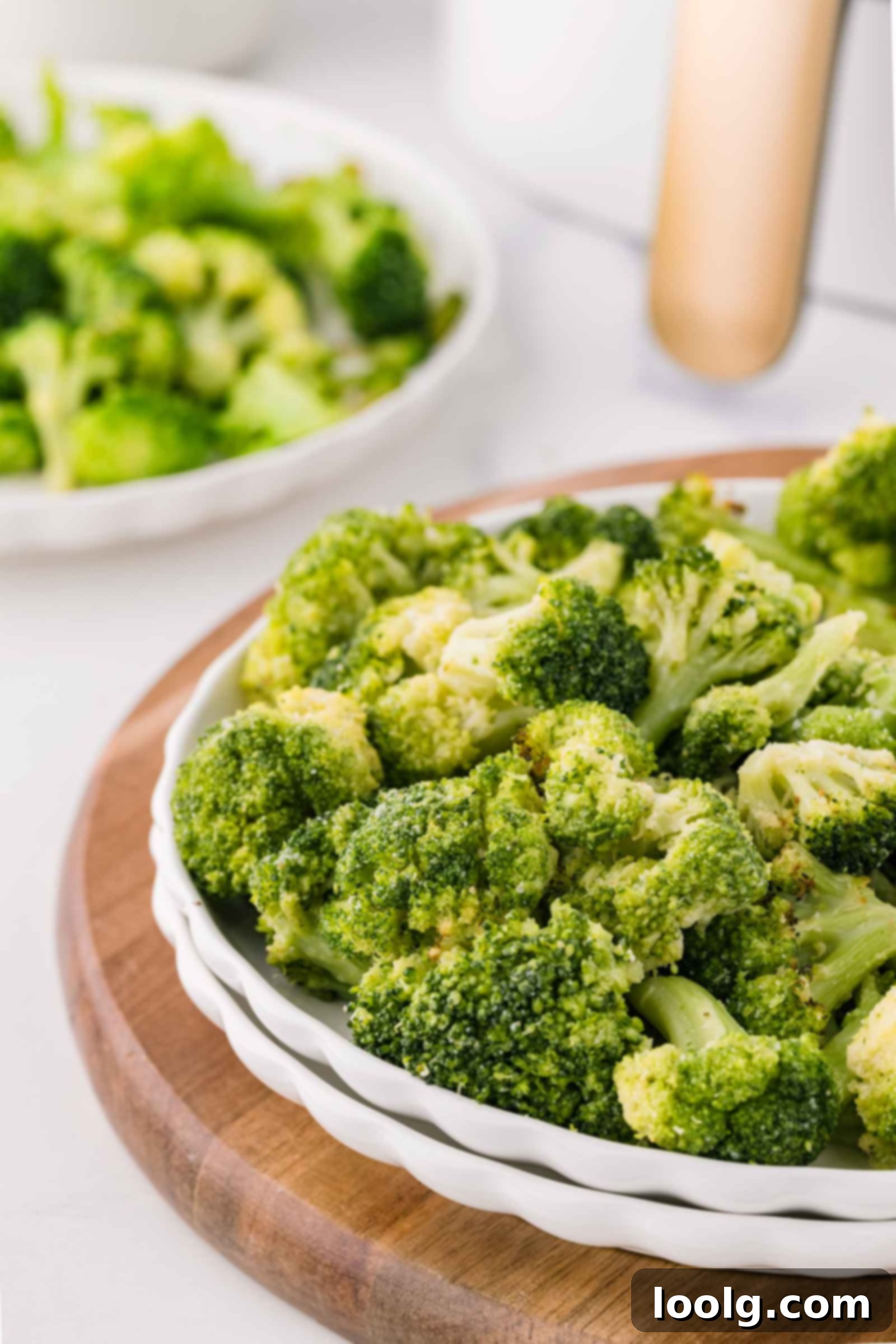 A white plate filled with air fryer frozen broccoli florets sits on a wooden board, with another dish of broccoli blurred in the background.