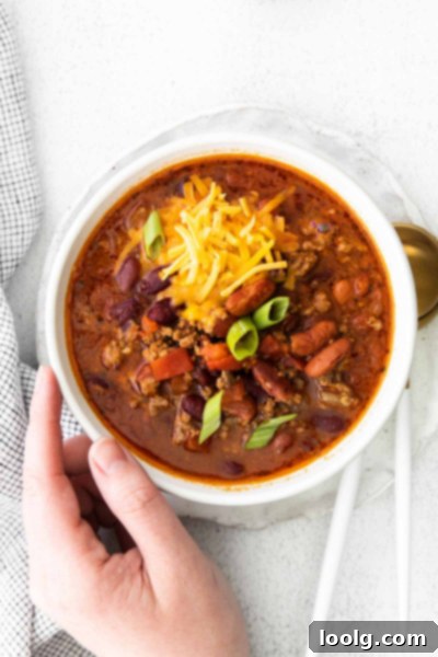 Venison Chili, Your Way: Slow Cooker or Stovetop 4 A close-up shot of a bowl of hearty venison chili, garnished with a sprig of fresh herb. The chili has a rich red-brown color with visible pieces of ground meat, beans, and diced tomatoes. The bowl is ceramic and sits on a rustic wooden surface, creating a warm, inviting feel.