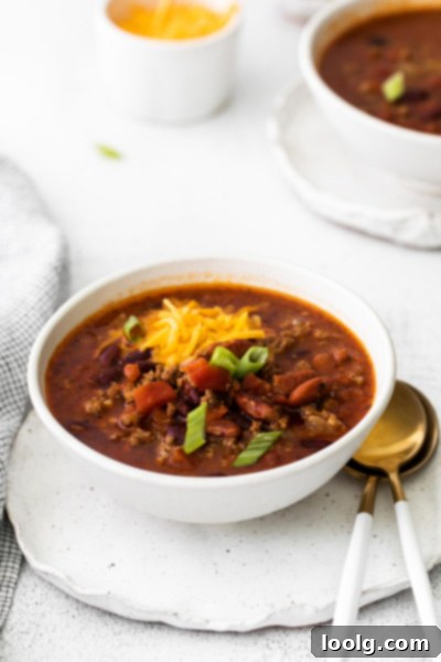 Venison Chili, Your Way: Slow Cooker or Stovetop 8 A vibrant bowl of venison chili sits on a table next to a sprig of fresh herb. The chili is steaming, indicating its warmth, and its rich color suggests deep flavor. The background is softly blurred, keeping the focus on the appetizing meal.