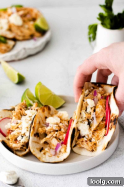 Effortless Instant Pot Shredded Chicken Tacos 2 Two shredded chicken tacos on a white plate with radish slices and lime wedges. One taco is being held by a hand.
