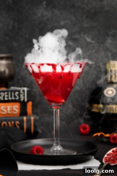 Crimson Kiss 2 A vibrant red Vampire's Kiss cocktail in a martini glass, naturally colored by raspberries and Chambord, with dry ice smoke swirling around it. Garnished with fresh raspberries and sitting on a dark plate, evoking a spooky Halloween atmosphere.