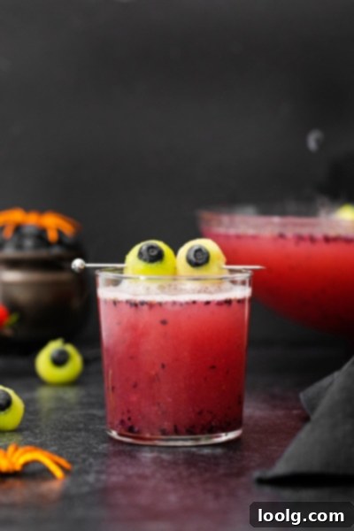 Fright Night Fizz 2 A glass of vibrant red non-alcoholic Halloween punch with floating melon and blueberry 'eyeballs' next to a spooky punch bowl.