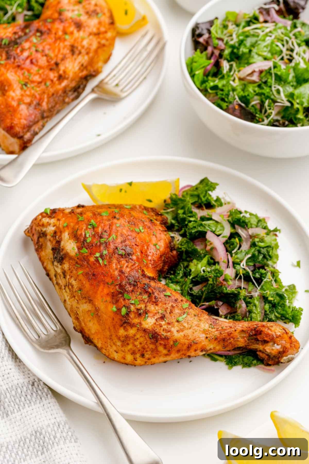 A golden-brown air fryer chicken quarter on a white plate, accompanied by a silver fork, a vibrant kale salad garnished with red onion slivers, and a fresh lemon wedge.