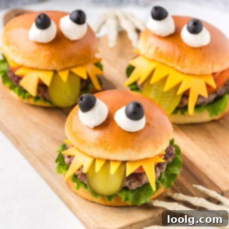 The Ultimate Monster Burger 15 image of three adorable-looking monster burgers for Halloween, each assembled to look like a monster with lettuce, tomatoes on the bottom bun, and a juicy hamburger patty. It looks like a monster with sharp teeth made of cheese, a long sliced green pickle tongue hanging out from under the top bun, and two eyes made of mozzarella balls and black olives secured by a toothpick on top of the brioche hamburger bun to make the burgers look like cute monsters.