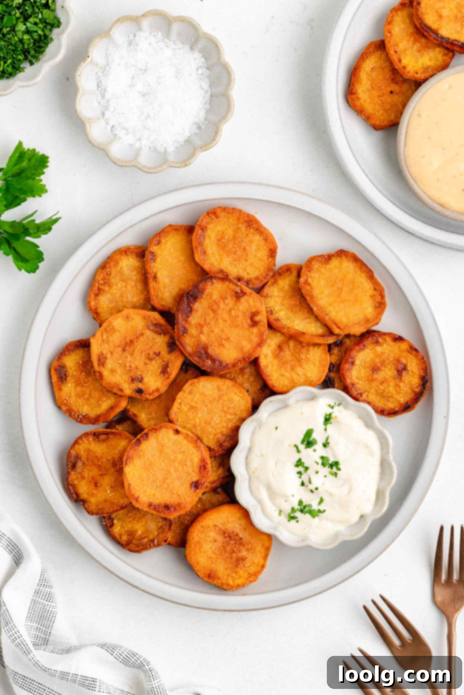 Golden Crispy Fried Sweet Potatoes 2 A plate of crispy fried sweet potatoes served with a small bowl of creamy dipping sauce, garnished with chopped herbs. Extra sauce, salt, and utensils are nearby on a white table.