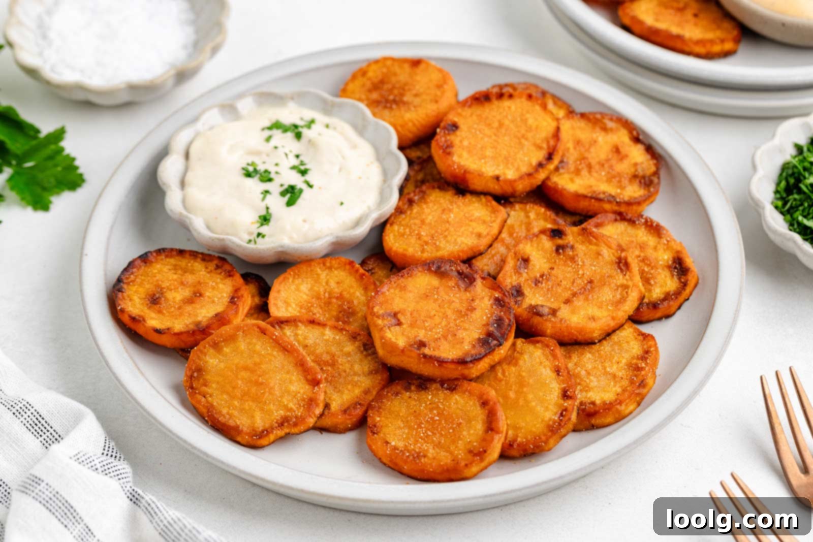 Golden Crispy Fried Sweet Potatoes 7 A plate of fried sweet potatoes is served with a small bowl of creamy dipping sauce topped with chopped herbs. A fork, napkin, and small bowls of salt and herbs are nearby.