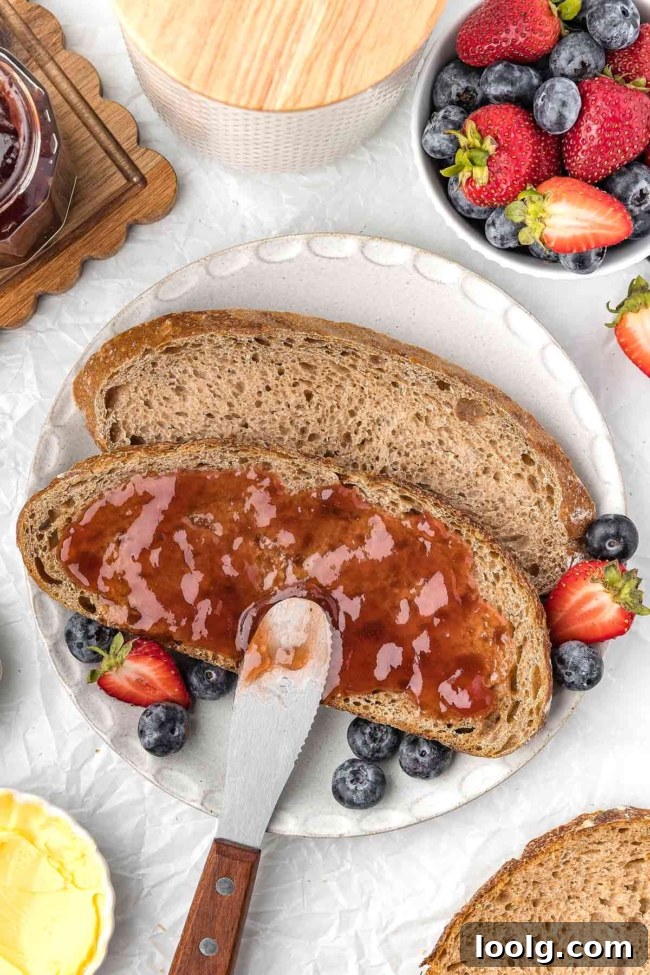 Whole wheat sourdough bread topped with jam, surrounded by fresh strawberries and blueberries on a plate.