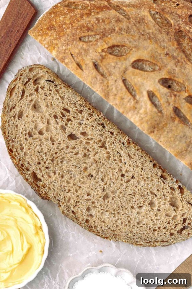 Whole wheat sourdough bread slice with a golden crust, accompanied by butter and salt on a textured surface.