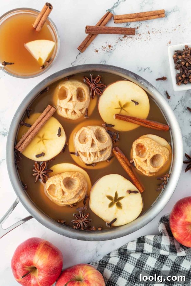 Shrunken Head Apple Cider: Spooky Halloween Drink Recipe 2 Spiced apple cider with carved apple shrunken heads, cinnamon sticks and star anise, surrounded by fresh apples and a checkered cloth.