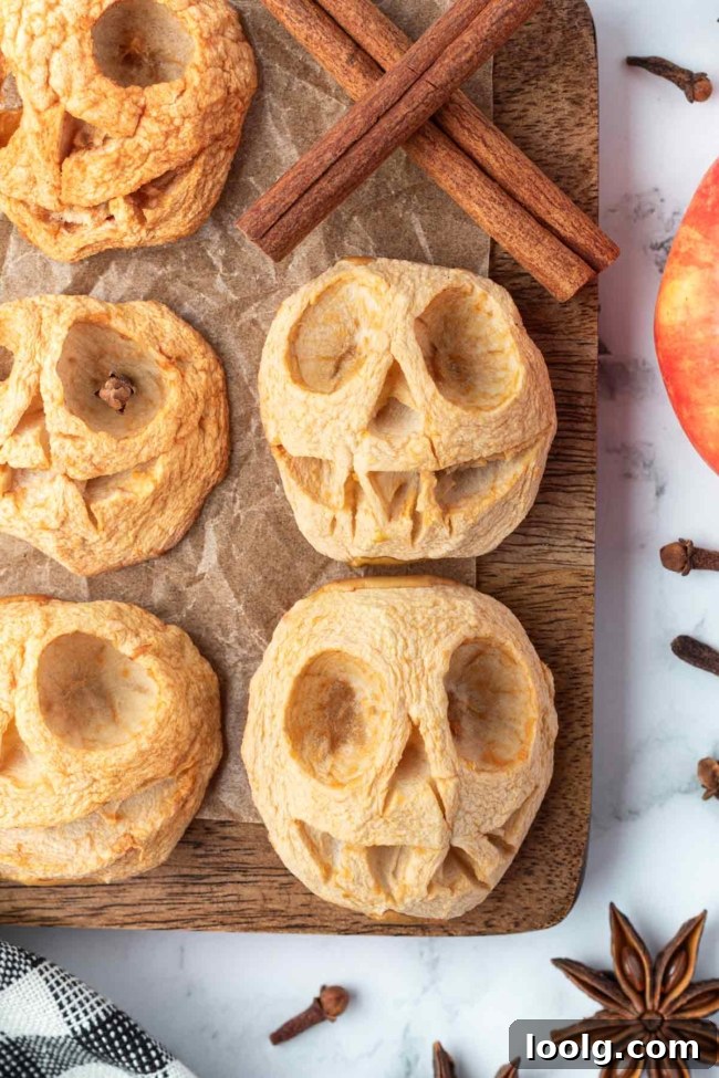 Shrunken Head Apple Cider: Spooky Halloween Drink Recipe 3 Carved apple halves baked into wrinkled shrunken heads, surrounded by cinnamon sticks and spices on a wooden board.